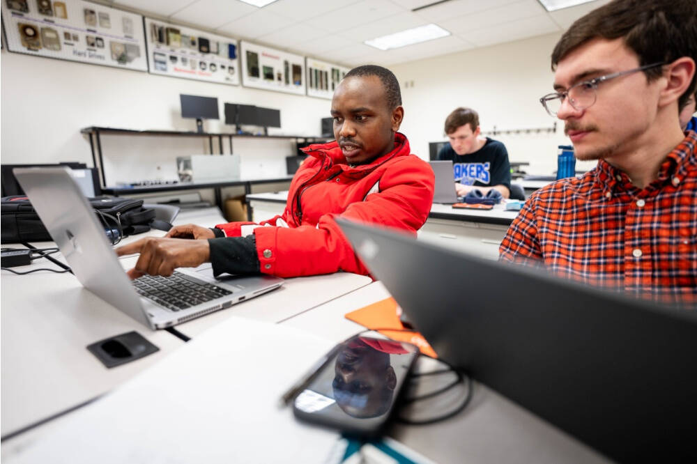 Two students in a classroom work together on laptops, with one in a red jacket pointing at the screen while the other observes, surrounded by electronic equipment and other focused classmates.